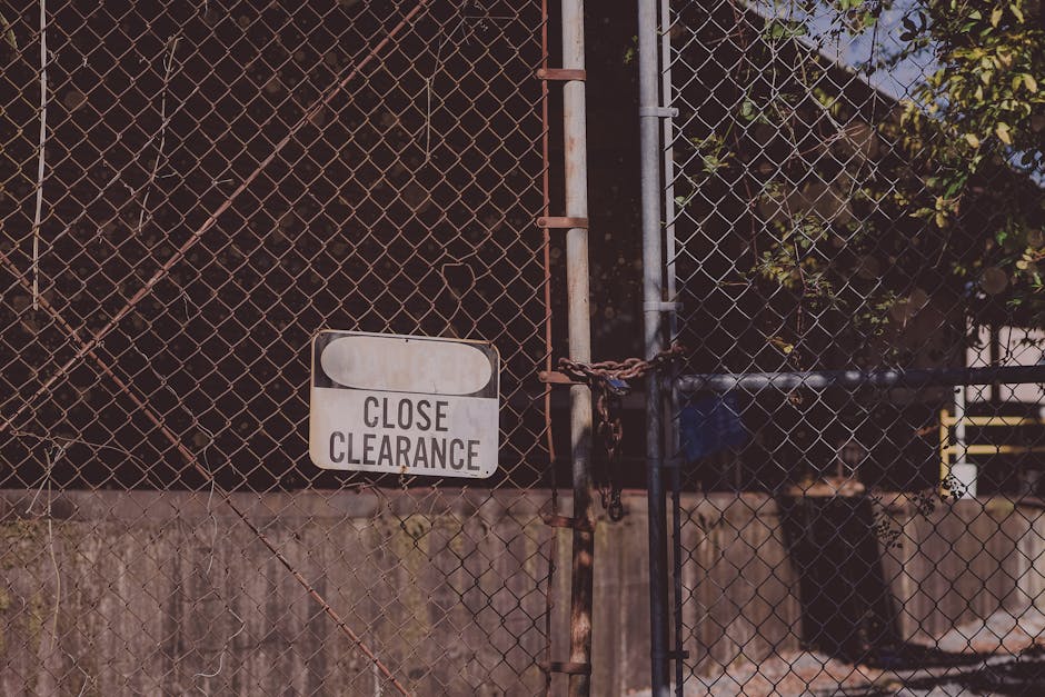 A close-up view of a metal garden gate made of chain-link fencing, secured with a rusty chain and padlock, preventing access to the area beyond. Attached to the gate is a small rectangular sign with r