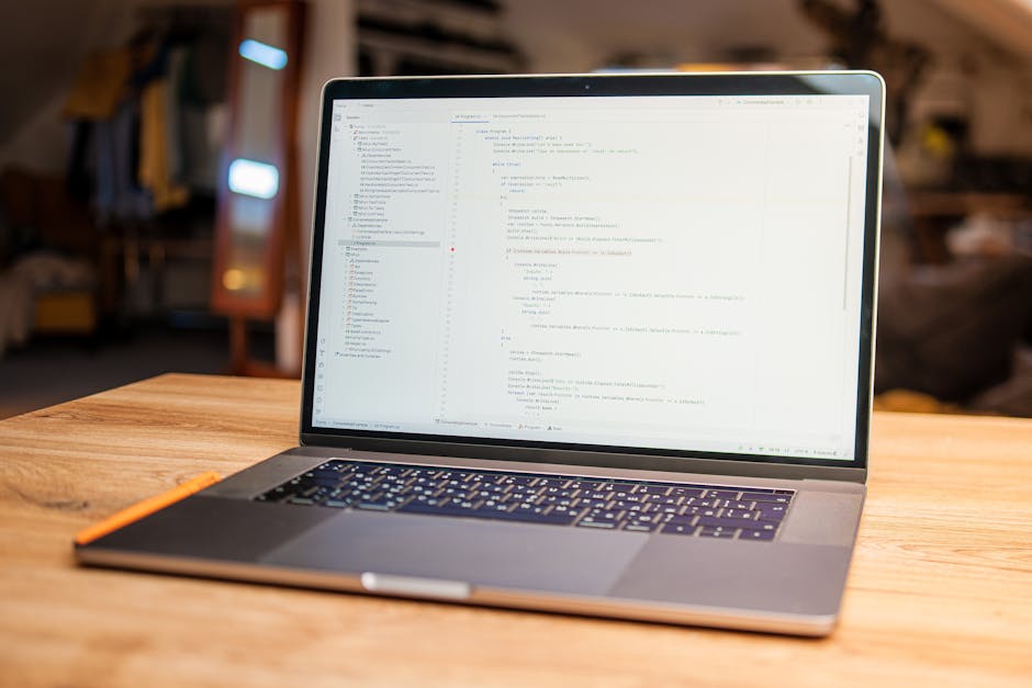 A silver MacBook laptop placed on a wooden table displaying lines of code on its screen, with an orange pencil resting alongside on the left. The background is indoors, featuring blurred furnishings a